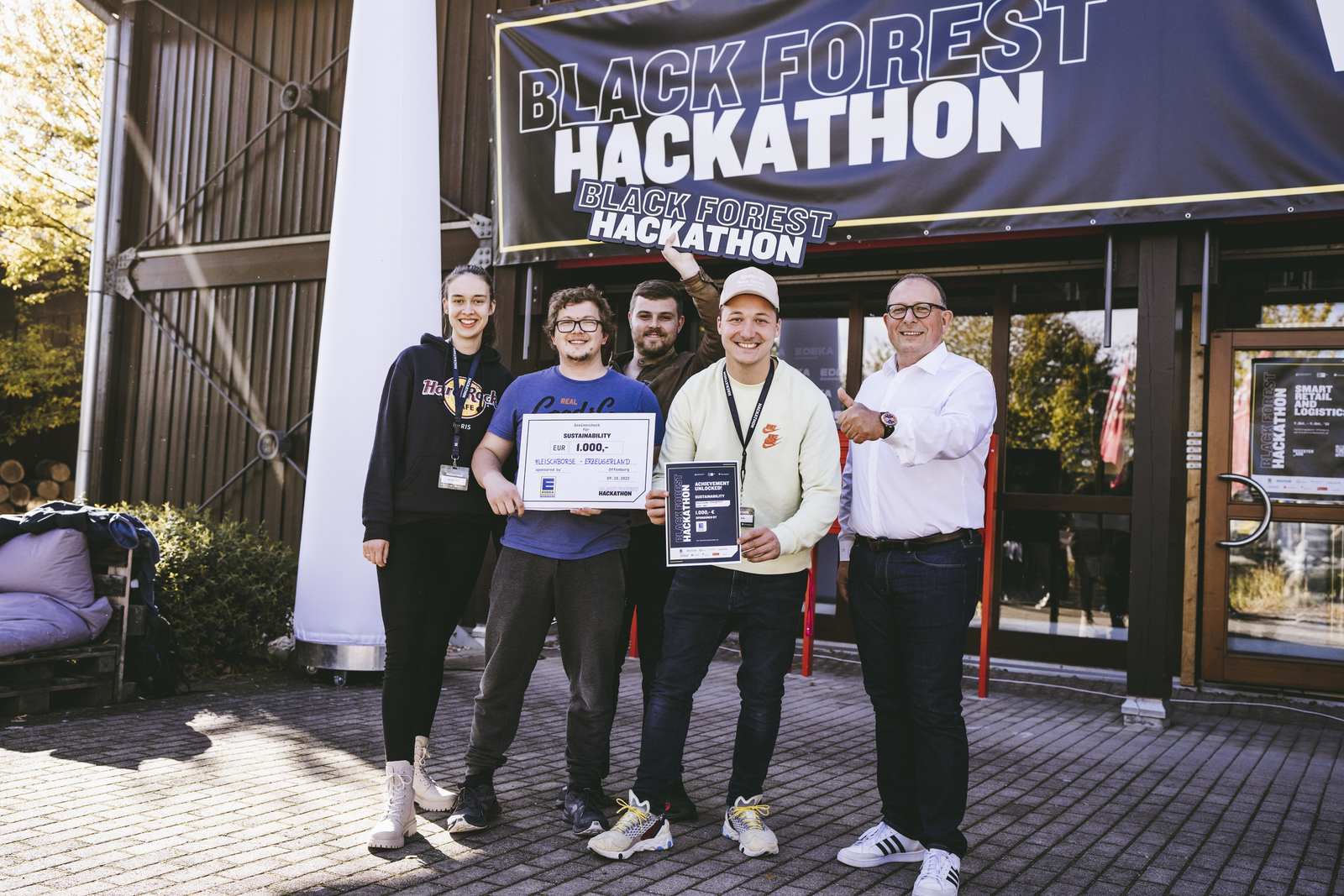 Das Gewinnerteam präsentiert vor einem Gebäude mit dem Banner Black Forest Hackathon seinen Siegerscheck und